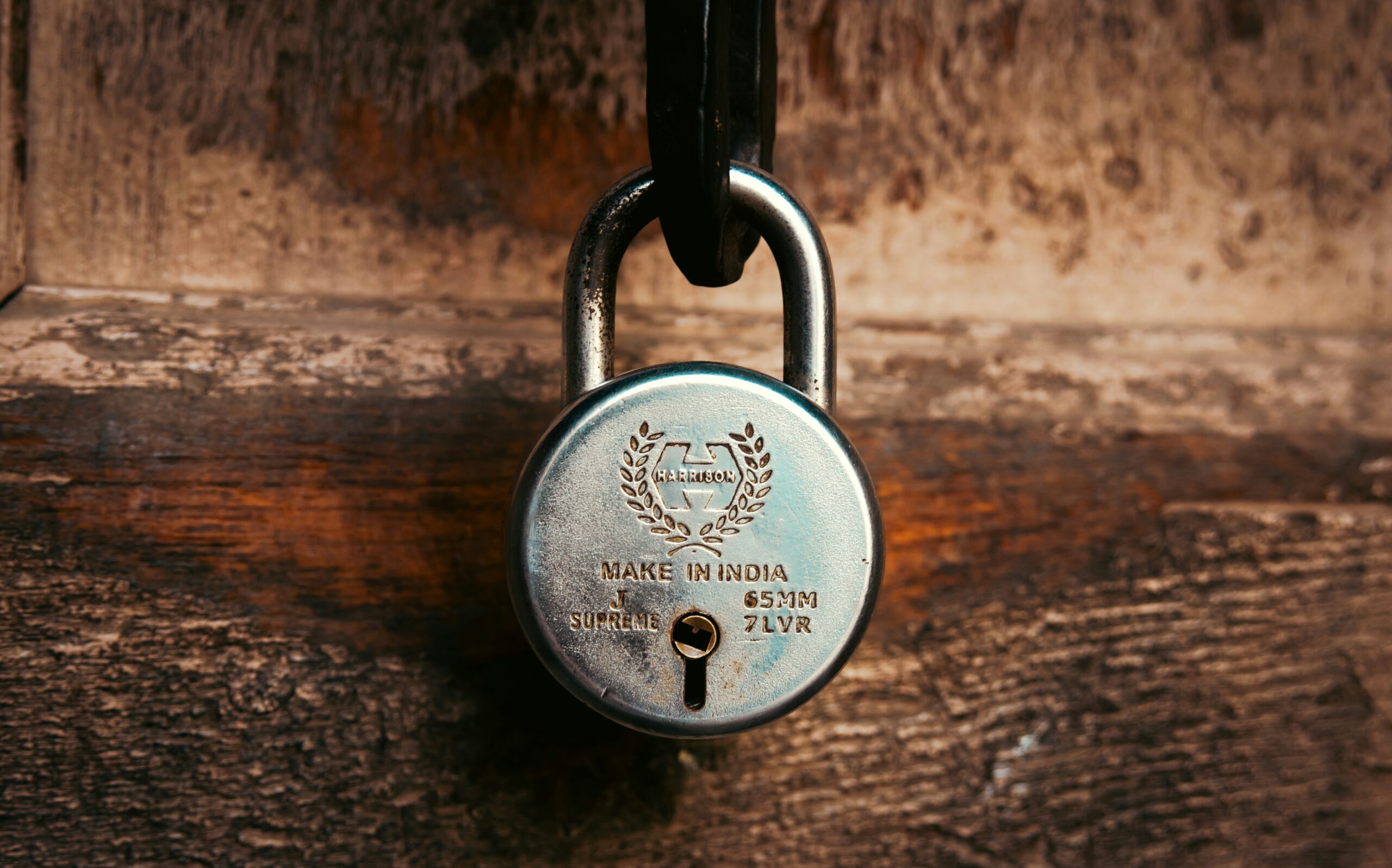 A close up view of a metal padlock hanging on an old wooden door. The lock, stamped with the Harrison brand and marked Make in India, stands out against the textured wood surface. The worn grain of the door and the polished finish of the lock create a strong contrast.